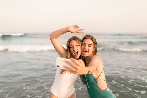 Dwa radosne żeńskie przyjaciółki biorąc selfie przed falami morza