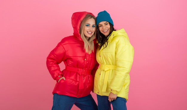 Dwie atrakcyjne dziewczyny pozują na różowym tle w kolorowej zimowej kurtce puchowej w jasnoczerwonym i żółtym kolorze