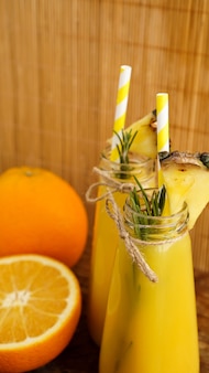 Dwie butelki soku tropikalnego z papierowymi słomkami. pomarańcze, ananas i rozmaryn do dekoracji. drewniane tło. zdjęcie pionowe