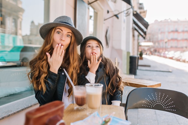 Dwie długowłose kręcone siostry patrzą na siebie z miłością, ciesząc się słonecznym porankiem w kawiarni na świeżym powietrzu.
