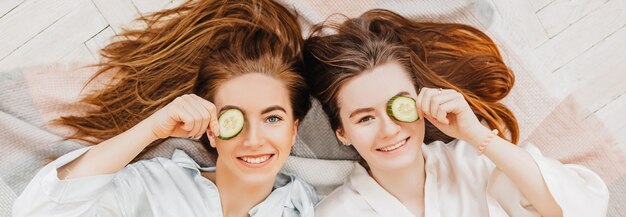 Dwie dziewczyny robią domowe maseczki do twarzy i włosów. ogórki dla świeżości skóry wokół oczu. kobiety dbają o młodzieńczą skórę. dziewczyny śmieją się w domu leżąc na podłodze na poduszkach.