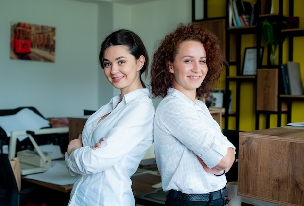 Bezpłatne zdjęcie dwie kobiety współpracowników biurowych patrząc na kamery z pewnym uśmiechem na twarzach stojących w biurze