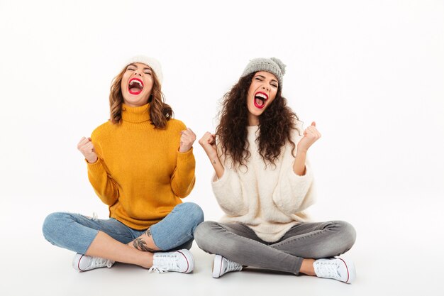 Dwie krzyczące dziewczyny w swetrach i kapeluszach siedzą razem na podłodze i radują się z białej ściany