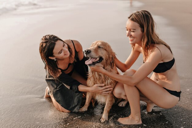 Dwie ładne dziewczyny na plaży w pobliżu oceanu, bawiąc się z psem na zachód słońca