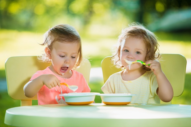 Dwie małe 2-letnie dziewczynki siedzące przy stole i jedzące razem na zielonym trawniku