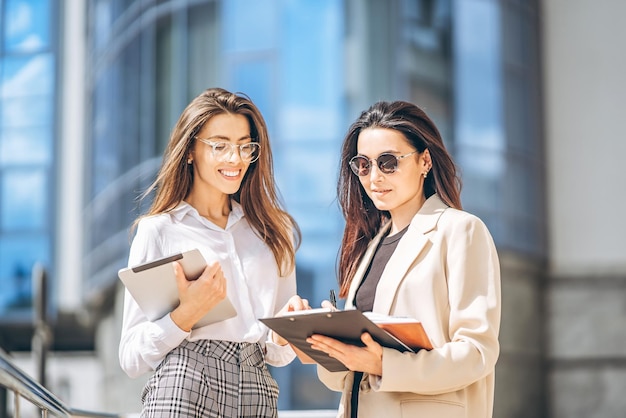 Dwie młode damy biznesu z tabletem i notebookiem spaceru na świeżym powietrzu w pobliżu nowoczesnego centrum biznesowego.