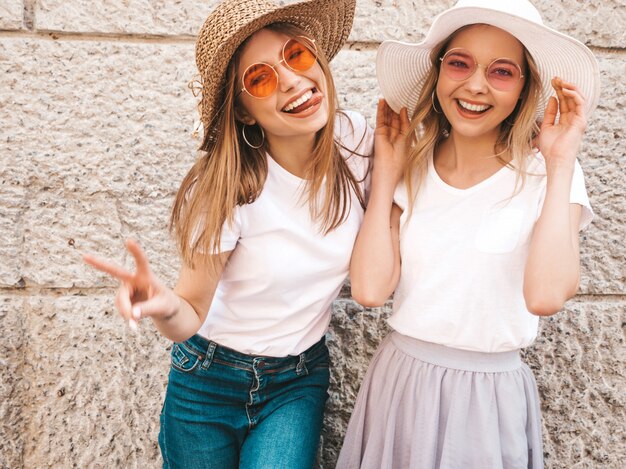Dwie młode piękne blond uśmiechnięte dziewczyny hipster w modne letnie białe ubrania t-shirt. Kobiety pozuje w ulicznej pobliskiej ścianie. . Pokazuje znak pokoju