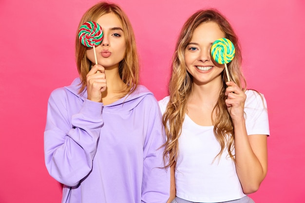 Bezpłatne zdjęcie dwie młode piękne uśmiechnięte blond hipster kobiety w modne letnie ubrania. beztroskie gorące kobiety pozuje blisko menchii ściany. pozytywne modele zakrywają oczy lollipop