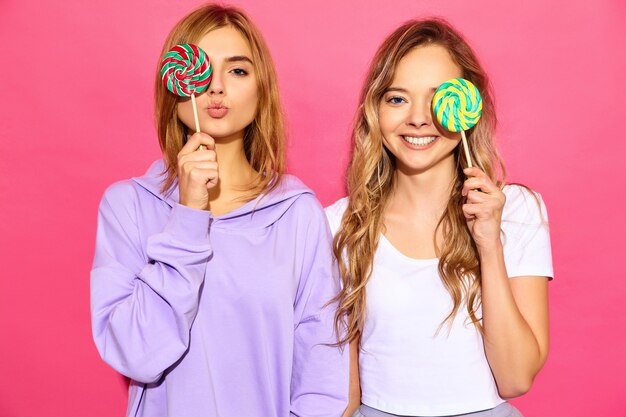 Dwie młode piękne uśmiechnięte blond hipster kobiety w modne letnie ubrania. Beztroskie gorące kobiety pozuje blisko menchii ściany. Pozytywne modele zakrywają oczy Lollipop
