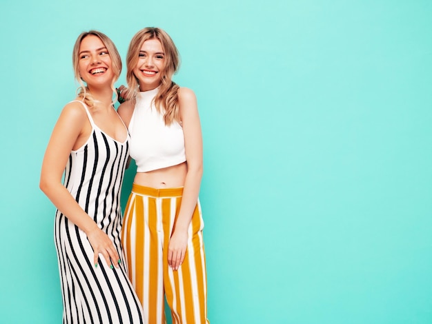 Dwie młode piękne uśmiechnięte brunetki hipster kobieta w modnych letnich ubraniach Seksowne beztroskie kobiety pozują w pobliżu niebieskiej ściany w studio Pozytywne modele zabawy Wesoły i szczęśliwy