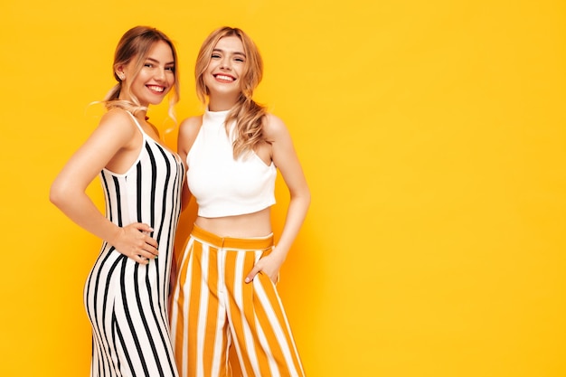Dwie młode piękne uśmiechnięte brunetki hipster kobieta w modnych letnich ubraniach Seksowne beztroskie kobiety pozują w pobliżu żółtej ściany w studio Pozytywne modele zabawy Wesoły i szczęśliwy