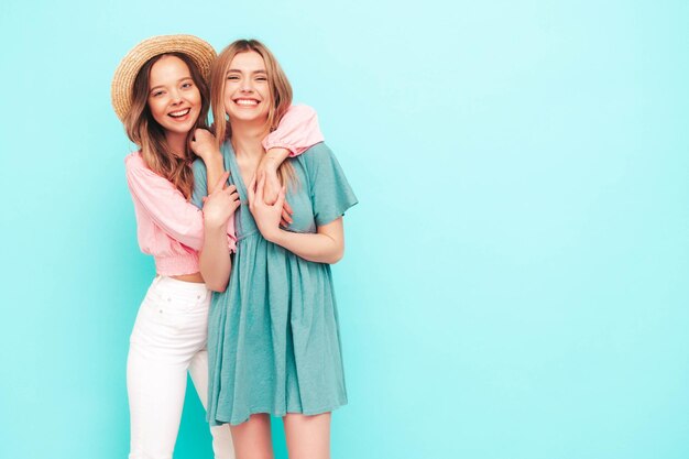 Dwie młode piękne uśmiechnięte brunetki hipster w modnych letnich sukienkach Seksowne beztroskie kobiety pozują w pobliżu niebieskiej ściany Pozytywne modele zabawy Wesoły i szczęśliwy w kapeluszach