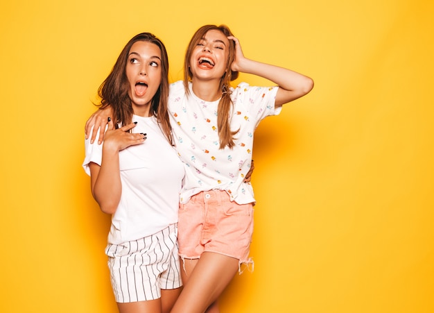 Dwie młode piękne uśmiechnięte dziewczyny hipster w modne letnie ubrania. Seksowne beztroskie kobiety pozuje blisko kolor żółty ściany. Pozytywne modele wariują i dobrze się bawią