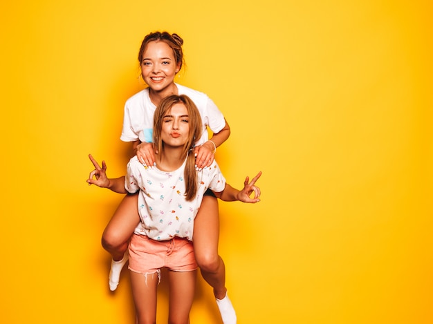 Dwie młode piękne uśmiechnięte dziewczyny hipster w modne letnie ubrania. Seksowne beztroskie kobiety pozuje blisko żółtej ściany Model siedzi na plecach przyjaciółki i pokazuje znak pokoju