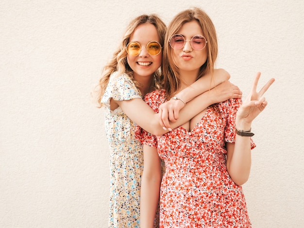 Dwie młode piękne uśmiechnięte hipster dziewczyny w modne letnie sukienki. Seksowne beztroskie kobiety pozowanie na ulicy w pobliżu ściany w okulary przeciwsłoneczne. Pozytywne modele zabawy i znak pokoju