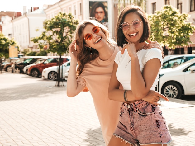 Dwie młode piękne uśmiechnięte hipster dziewczyny w modne letnie ubrania. Seksowne beztroskie kobiety pozowanie na tle ulicy w okulary przeciwsłoneczne. Pozytywne modele zabawy i szaleństwa