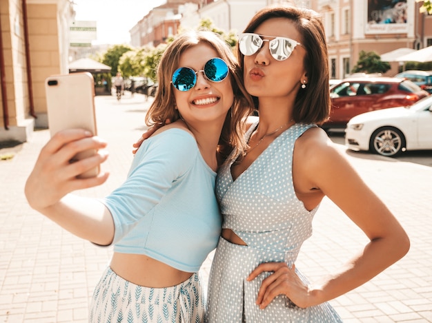 Dwie młode piękne uśmiechnięte hipster dziewczyny w modne letnie ubrania. Seksowne beztroskie kobiety pozowanie na tle ulicy w okulary przeciwsłoneczne. Robią selfie autoportrety na smartfonie o zachodzie słońca