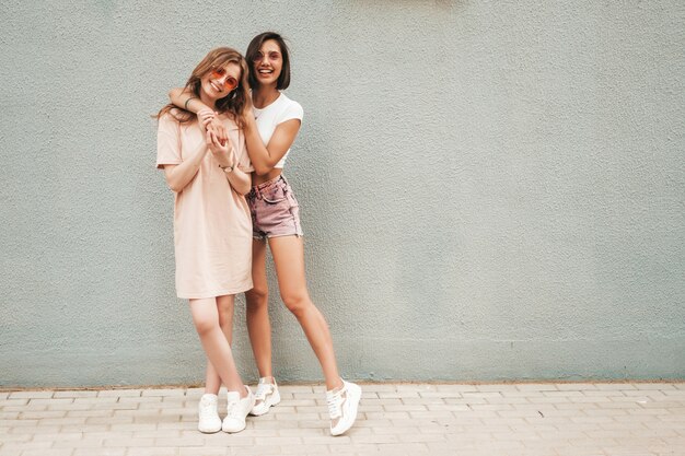 Dwie młode piękne uśmiechnięte hipster dziewczyny w modne letnie ubrania. Seksowne beztroskie kobiety pozuje na ulicznym tle w okularach przeciwsłonecznych. Pozytywne modele zabawy i przytulania