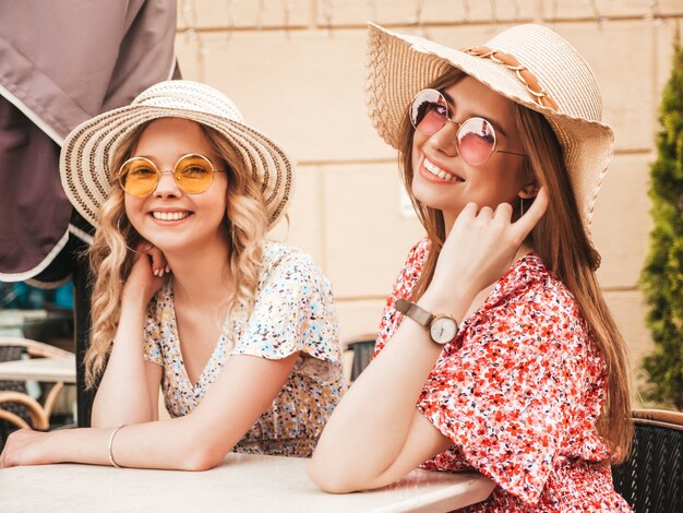Dwie młode piękne uśmiechnięte hipster dziewczyny w modnej letniej sukience. Beztroskie kobiety na czacie w kawiarni werandzie na tle ulicy w okularach przeciwsłonecznych. Pozytywne modele zabawy i komunikacji