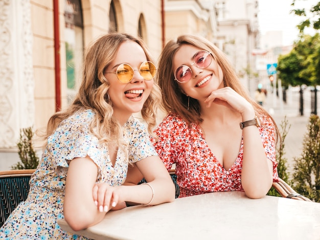 Dwie młode piękne uśmiechnięte hipster dziewczyny w modnej letniej sukience. Beztroskie kobiety na czacie w kawiarni werandzie na tle ulicy w okularach przeciwsłonecznych. Pozytywne modele zabawy i komunikacji
