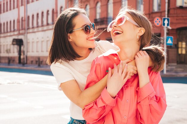 Dwie młode piękne uśmiechnięte hipster kobieta w modne letnie ubrania i dżinsy biały t-shirt. Seksowne beztroskie kobiety pozowanie na tle ulicy. Pozytywne modelki bawią się, przytulają i szaleją