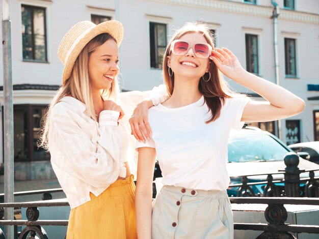 Dwie młode piękne uśmiechnięte kobiety hipster w modnych letnich ubraniachSeksowne beztroskie kobiety pozowanie na tle ulicy w kapeluszu Pozytywne czyste modele zabawy o zachodzie słońca przytulanie