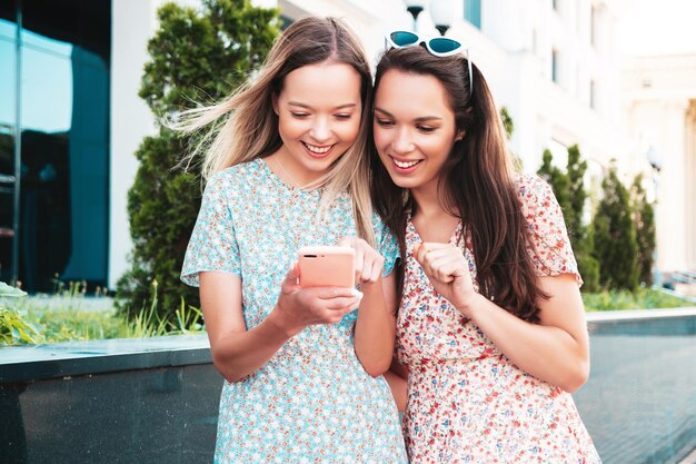 Dwie młode piękne uśmiechnięte kobiety hipster w modnych letnich ubraniachSeksowne beztroskie kobiety pozują na ulicy Pozytywne modele zabawy przytulanie Patrzą na ekran smartfona za pomocą aplikacji na telefon