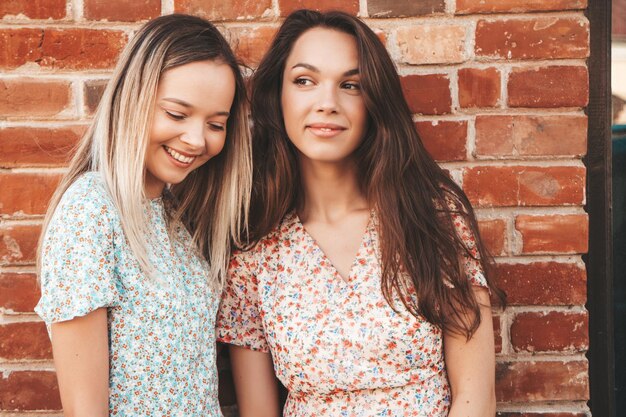 Dwie młode piękne uśmiechnięte kobiety hipster w modnych letnich ubraniachSeksowne beztroskie kobiety pozują na ulicy w pobliżu ceglanego muru Pozytywne czyste modele bawiące się o zachodzie słońca Szczęśliwe i wesołe
