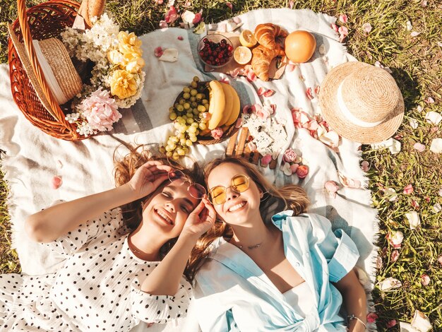 Dwie młode piękne uśmiechnięte kobiety w modnej letniej sukience i kapeluszach. Beztroskie kobiety robią piknik na zewnątrz.