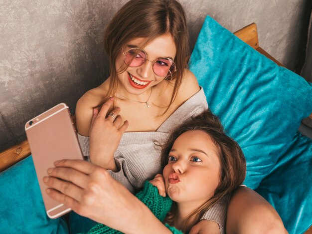 Dwie młode piękne uśmiechnięte wspaniałe dziewczyny w modne letnie ubrania. Seksowne beztroskie kobiety pozuje w wnętrzu i bierze selfie. Pozytywne modele zabawy ze smartfonem.