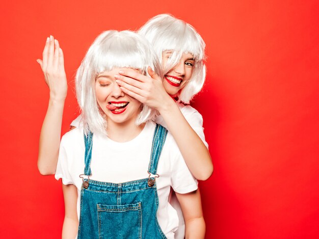 Dwie młode seksowne uśmiechnięte dziewczyny hipster w białe peruki i czerwone usta. Piękne modne kobiety w letnie ubrania. Modele pozowanie w pobliżu czerwonej ściany w studio. Zasłonić oczy dłońmi do swojej przyjaciółki. Koncepcja niespodzianka