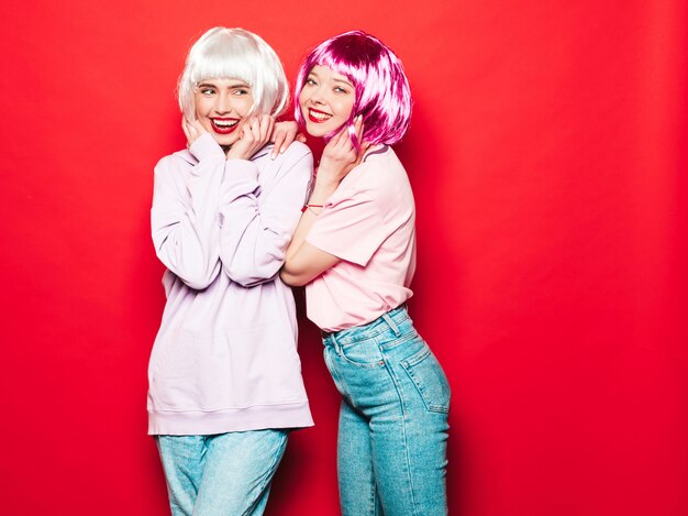 Dwie młode seksowne uśmiechnięte hipster dziewczyny w białych perukach i czerwonych ustach. Piękne modne kobiety w letnie ubrania. Beztroskie modele pozowanie w pobliżu czerwonej ściany w studio wariuje