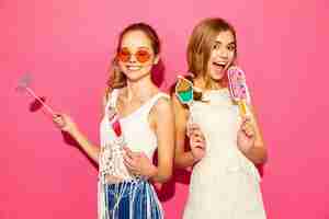 Bezpłatne zdjęcie dwie młode stylowe uśmiechnięte blond kobiety jedzące rekwizyty słodkie lody i fałszywy koktajl. pozytywne modele w letnich ubraniach hipster pozowanie w pobliżu różowej ściany w okularach przeciwsłonecznych