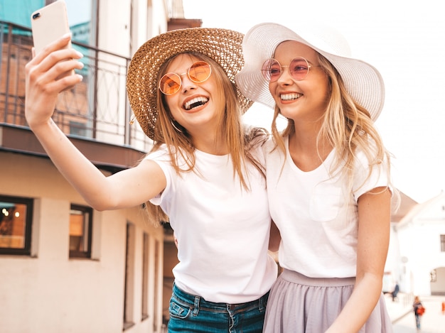 Dwie młode uśmiechnięte hipster blond kobiety w letnie białe ubrania ubrania. Dziewczyny robienia zdjęć autoportretów selfie na smartfonie. .