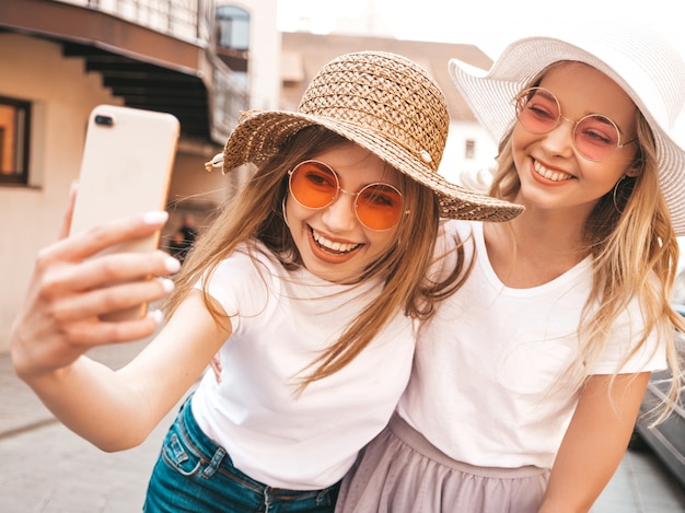 Bezpłatne zdjęcie dwie młode uśmiechnięte hipster blond kobiety w letnie białe ubrania ubrania. dziewczyny robienia zdjęć autoportretów selfie na smartfonie. .