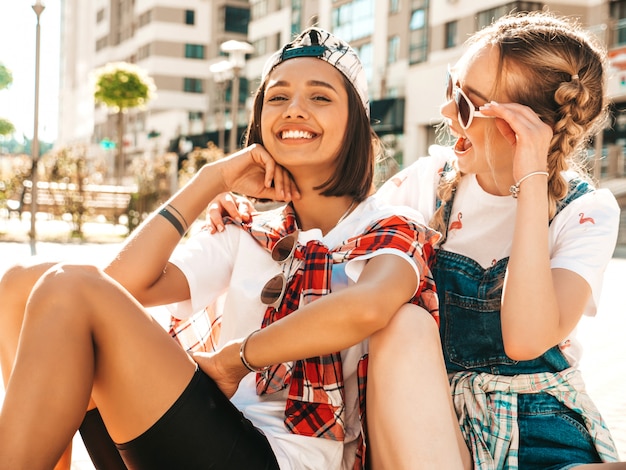 Bezpłatne zdjęcie dwie młode uśmiechnięte piękne dziewczyny z kolorowymi deskorolkami grosza. kobiety w letnie ubrania hipster siedzi na tle ulicy. pozytywne modelki bawią się i wariują