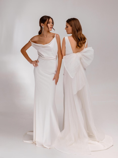 Dwie modne kobiety brunetki w sukniach ślubnych i fryzurach ślubnych stojących razem pozowanie na białym tle Urocze panny młode Koncepcja strzyżenia biżuterii ślubnej Salon ślubny