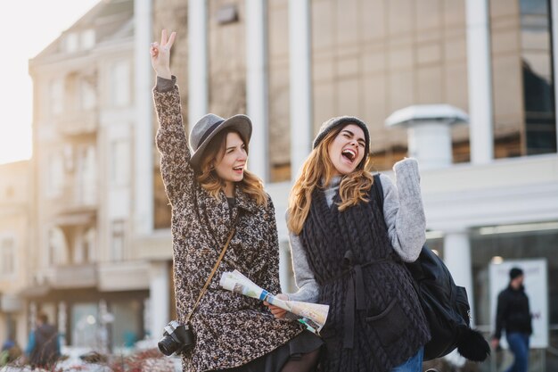 Dwie modne, radosne uśmiechnięte kobiety skaczące po mieście. Stylowy wygląd, wspólne podróżowanie, noszenie modnych ubrań, spacery z kawą na wynos, wyrażanie pozytywnych emocji.