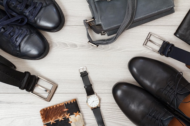 Dwie pary butów męskich z czarnej skóry, dwa paski męskie, torba na ramię, portfel męski, zegarek na rękę i torebka na szarych drewnianych deskach. akcesoria męskie