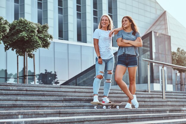 Dwie piękne dziewczyny hipster stojące na schodach z deskorolką na tle wieżowca.