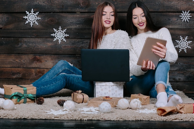 Dwie piękne kobiety siedzące na podłodze z laptopem i tabletem, między prezentami na Boże Narodzenie