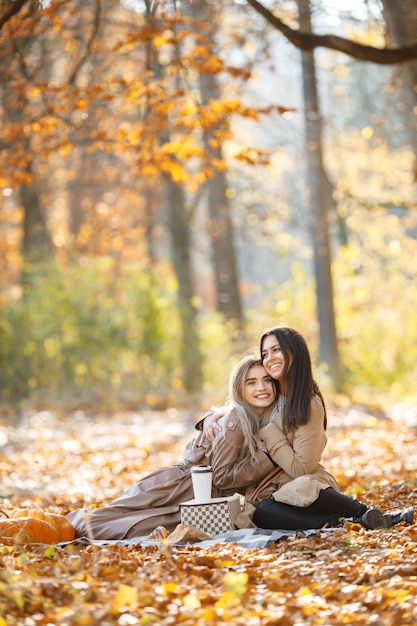 Dwie piękne koleżanki spędzają czas na kocu piknikowym na trawie. Dwie młode uśmiechnięte siostry robią piknik jedząc rogalika w jesiennym parku. Brunetki i blondynki w płaszczach.