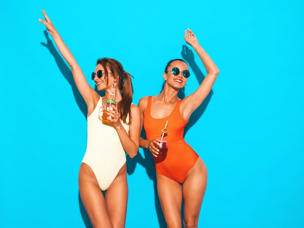 Dwie piękne seksowne uśmiechnięte kobiety w kolorowe kolorowe stroje kąpielowe kostiumy kąpielowe. Modne dziewczyny w okularach przeciwsłonecznych. Wariować. Śmieszne modele na białym tle. Picie świeżego smoozy drinka koktajlowego. Podnieść ręce