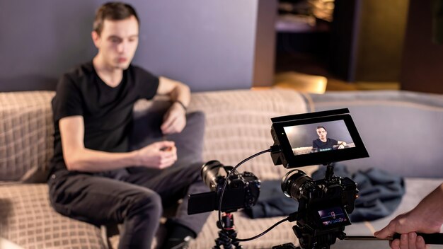 Dwie profesjonalne kamery wideo na statywie rejestrujące rozmawiającego mężczyznę siedzącego na kanapie w domu. Praca z domu