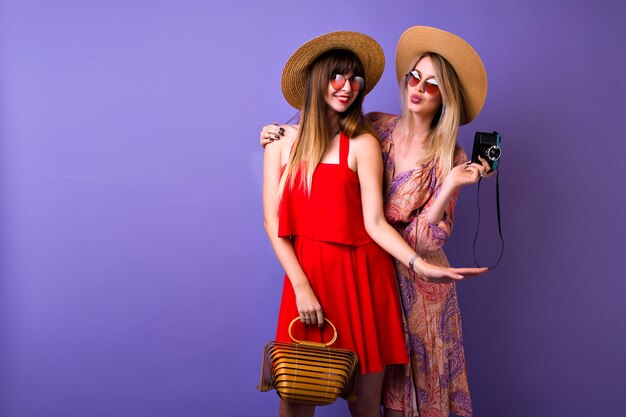 Dwie stylowe hipster dziewczyny bawiące się razem, czapki i akcesoria do sukienki w stylu boho w stylu vintage, blondynka robi zdjęcia swojej najlepszej przyjaciółce,