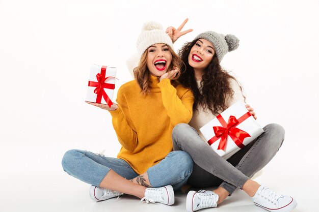 Dwie szczęśliwe dziewczyny w swetrach i czapkach, siedzące razem z prezentami na podłodze podczas zabawy nad białą ścianą
