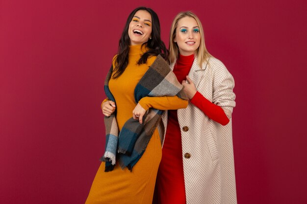 Dwie uśmiechnięte atrakcyjne stylowe kobiety w jesień zima moda sukienka i płaszcz pozowanie na białym tle na czerwonej ścianie