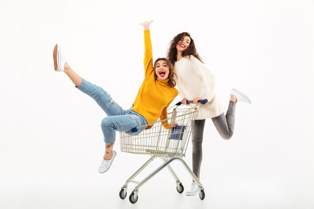 Dwie wesołe dziewczyny w swetrach bawiących się razem z wózkiem na zakupy