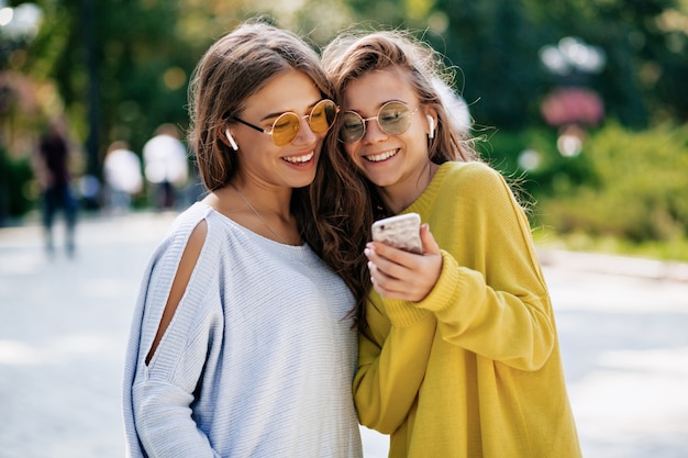 Dwie zabawnie uśmiechnięte siostry robiące selfie na smaptphone i słuchające muzyki, pozujące na ulicy, wakacyjny nastrój, szalone pozytywne uczucie, letnie jasne okulary przeciwsłoneczne.