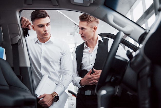 Bezpłatne zdjęcie dwóch mężczyzn stoi w salonie przed samochodami. zbliżenie na kierownika sprzedaży w garniturze, który sprzedaje samochód klientowi. sprzedawca przekazuje klucz klientowi.